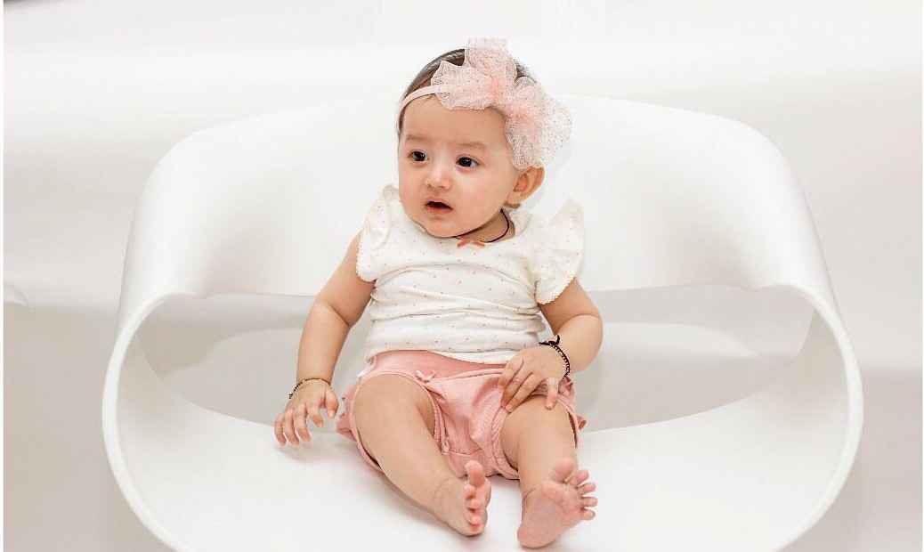 Baby girl sitting like a little angel in a white dress in a charming baby photoshoot.